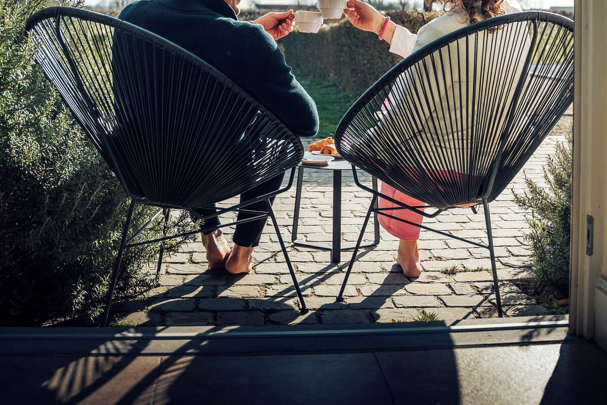 Two people sitting on outdoor chairs facing each other, holding cups, on a patio with a clear sky.