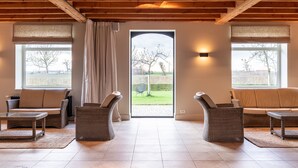 Ein geräumiger Raum mit Holzbalkendecke, zwei Sofas, Korbsesseln, einem großen Fenster und Blick auf ein Wiesfeld.