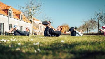 Een groep mensen ligt op het gras in een openlucht omgeving, met gebouwen op de achtergrond.