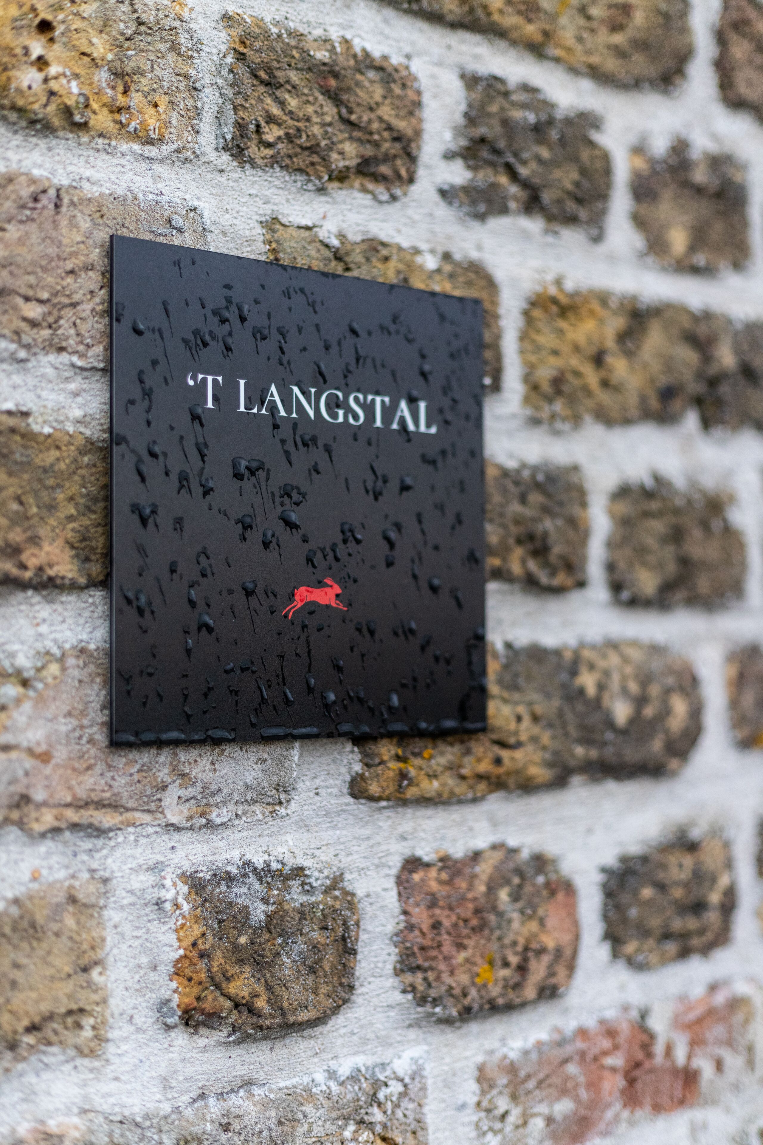 A black sign with the text "T. Langstal" and a red deer symbol on a brick wall.