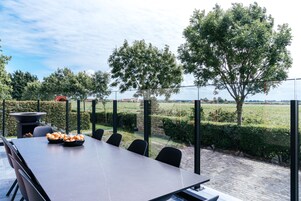 An outdoor dining area with a table and chairs, a bowl of bread, and a view of a landscaped garden and fields.