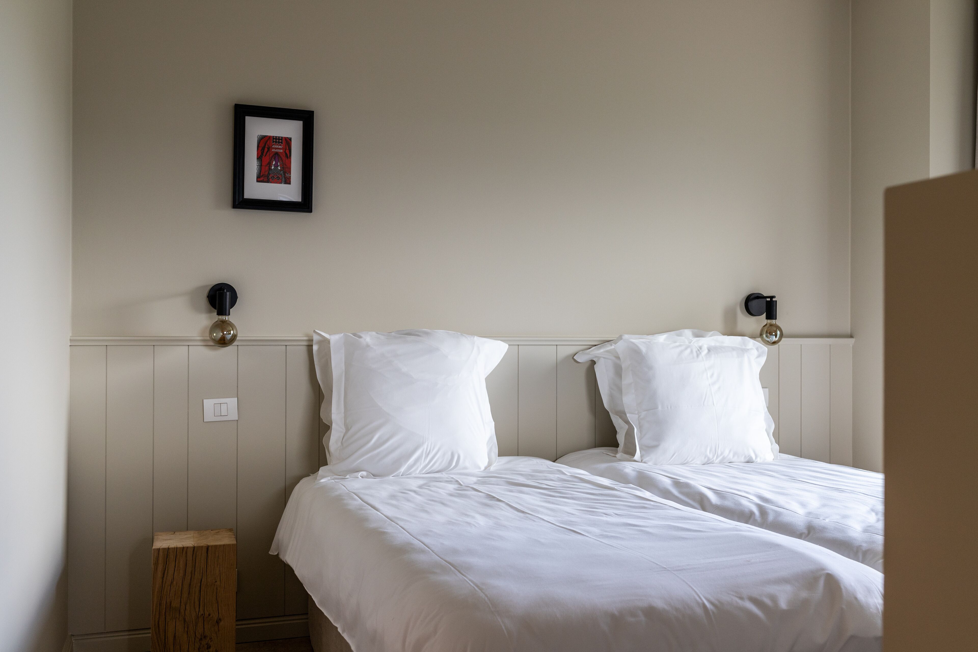 Una cama doble con ropa blanca, un taburete de madera y un cuadro en la pared.