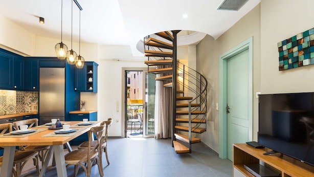 Une cuisine moderne avec des meubles bleus, un coin repas avec une table et des chaises en bois, un escalier en colimaçon et un téléviseur posé sur un meuble en bois.