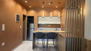 A compact kitchen with a refrigerator, two bar stools, and a wooden partition. - Deluxe Studio – Comfort & Style for Two (Crete)