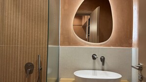 A modern bathroom with a glass shower enclosure, a wooden vanity with a white sink, and a large oval mirror. - Deluxe Studio – Comfort & Style for Two (Crete)
