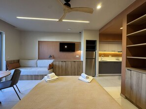 A modern hotel room with a bed, a desk, a kitchenette, and a TV mounted on the wall.