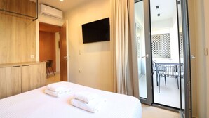 Ein Hotelzimmer mit Bett, einem an der Wand montierten Fernseher, einer Schiebetür zu einem Balkon mit Tisch und Stühlen sowie einer Klimaanlage an der Wand.