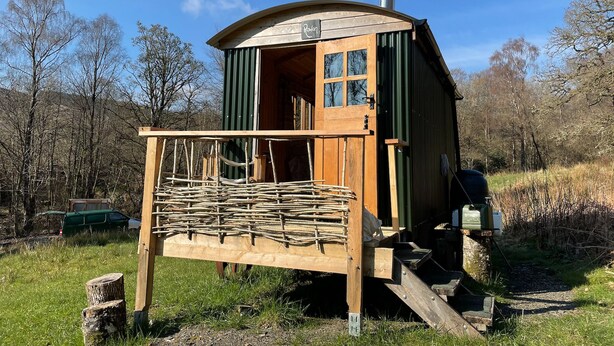 A wooden cabin with a chimney, a porch with wooden railings, and a door with windows. - Cosy Woodland off Grid Shepherds Hut - Rowan (Castle Douglas)