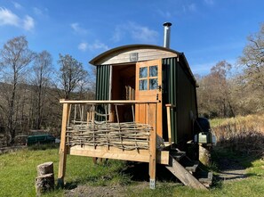 A wooden cabin with a chimney, a porch with wooden railings, and a door with windows. - Cosy Woodland off Grid Shepherds Hut - Rowan (Castle Douglas)