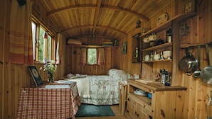 A cozy cabin interior with a bed, a table with a checkered tablecloth, a window with curtains, and wooden shelves with kitchenware. - Cosy Woodland off Grid Shepherds Hut - Rowan (Castle Douglas)