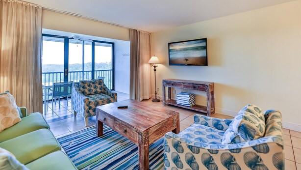 A living room with a sofa, a coffee table, a TV, a fireplace, and a balcony with a view. - SOUTH Villa 5310 - Three bedroom condo with Bay views (Captiva)
