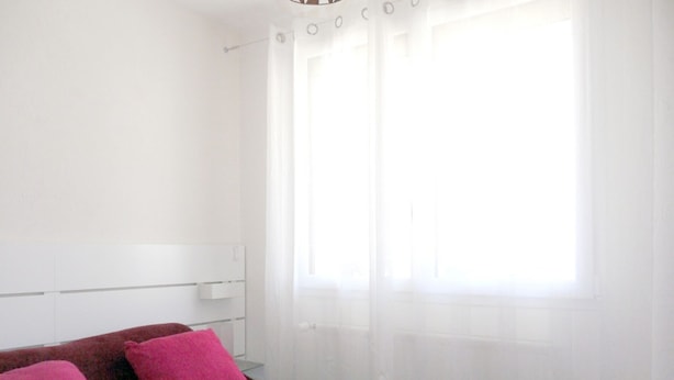 Ein Schlafzimmer mit einem Bett, zwei rosafarbenen Kissen, einem weißen Kopfstück und einem Fenster mit durchsichtigen Vorhängen.