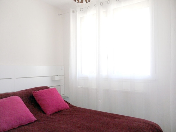 Ein Schlafzimmer mit einem Bett, zwei rosafarbenen Kissen, einem weißen Kopfstück und einem Fenster mit durchsichtigen Vorhängen.