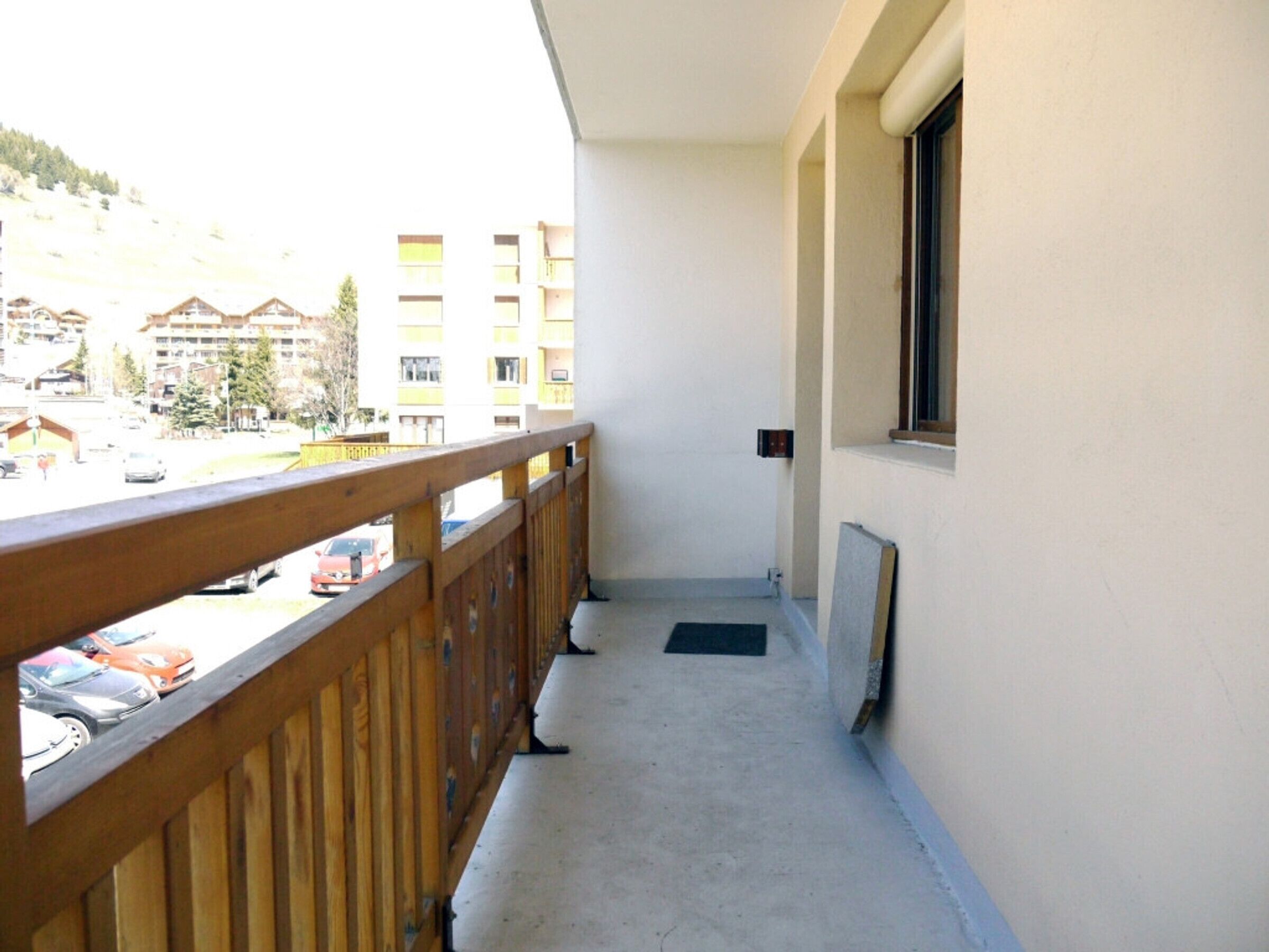 Un balcón con barandilla de madera, una ventana y vista a un estacionamiento y edificios al fondo.