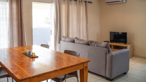 A living room with a wooden dining table, grey sofa, and a TV on a wooden stand. - Dei Gratia Guesthouse (Durban)