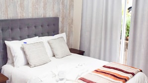 A neatly made bed with a striped blanket, a tufted headboard, and a view of a garden through the window. - Dei Gratia Guesthouse (Durban)