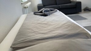 Una cama con una manta doblada y una almohada, un sofá gris, un televisor montado en la pared y una planta colgando de la pared.
