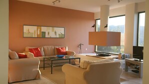 A modern living room with a sofa, armchairs, a coffee table, and a television. There is a large window providing natural light and a view of the outdoors. - Lovely Open Plan Family Villa in Kato Katelios (Kefalonia)