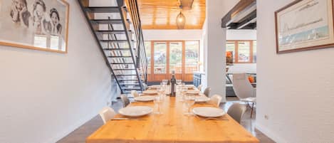 Un comedor con una larga mesa de madera dispuesta para una comida, una escalera y cuadros en las paredes.
