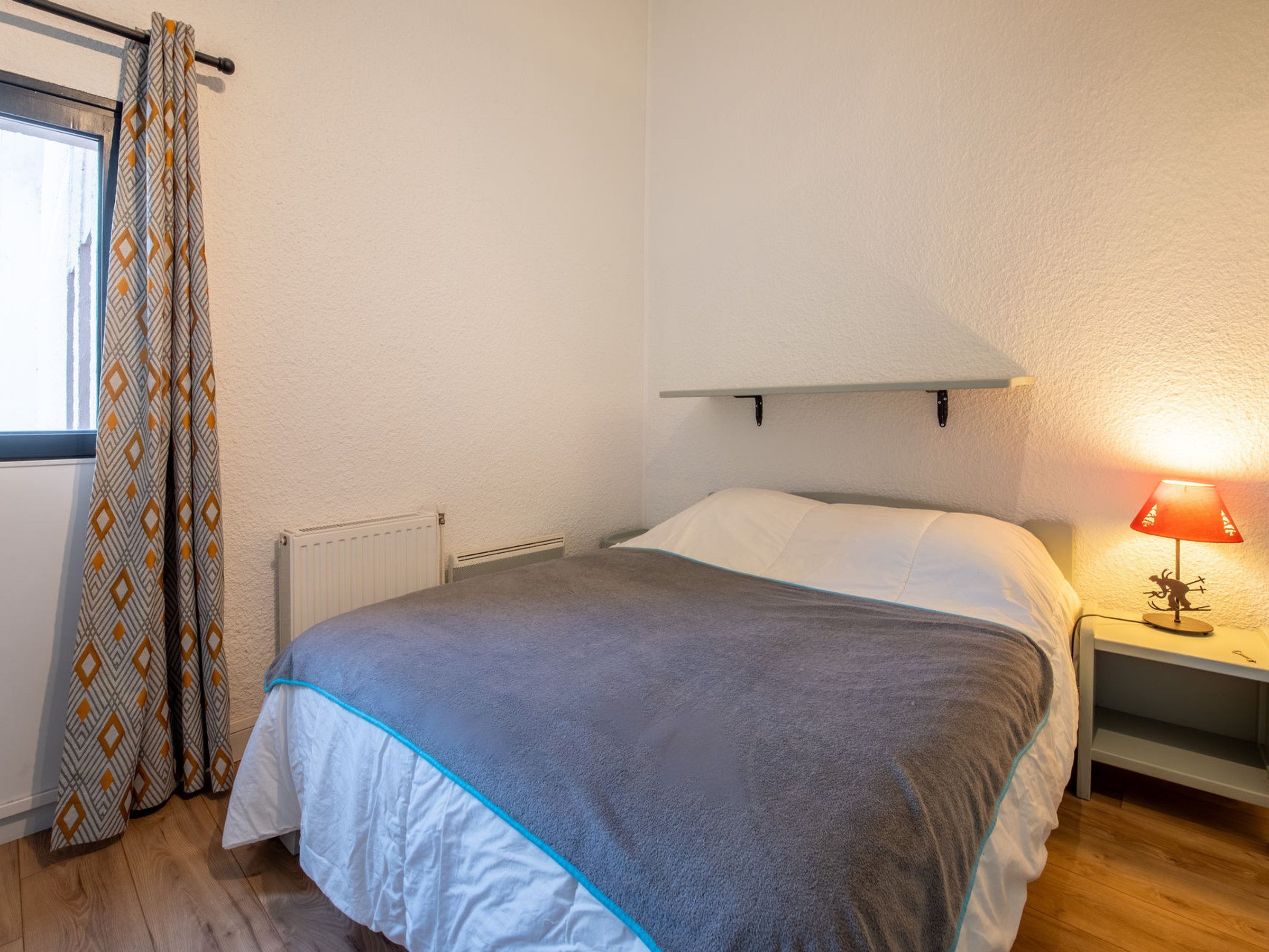 A bedroom with a bed, a nightstand with a lamp, a radiator, and patterned curtains.