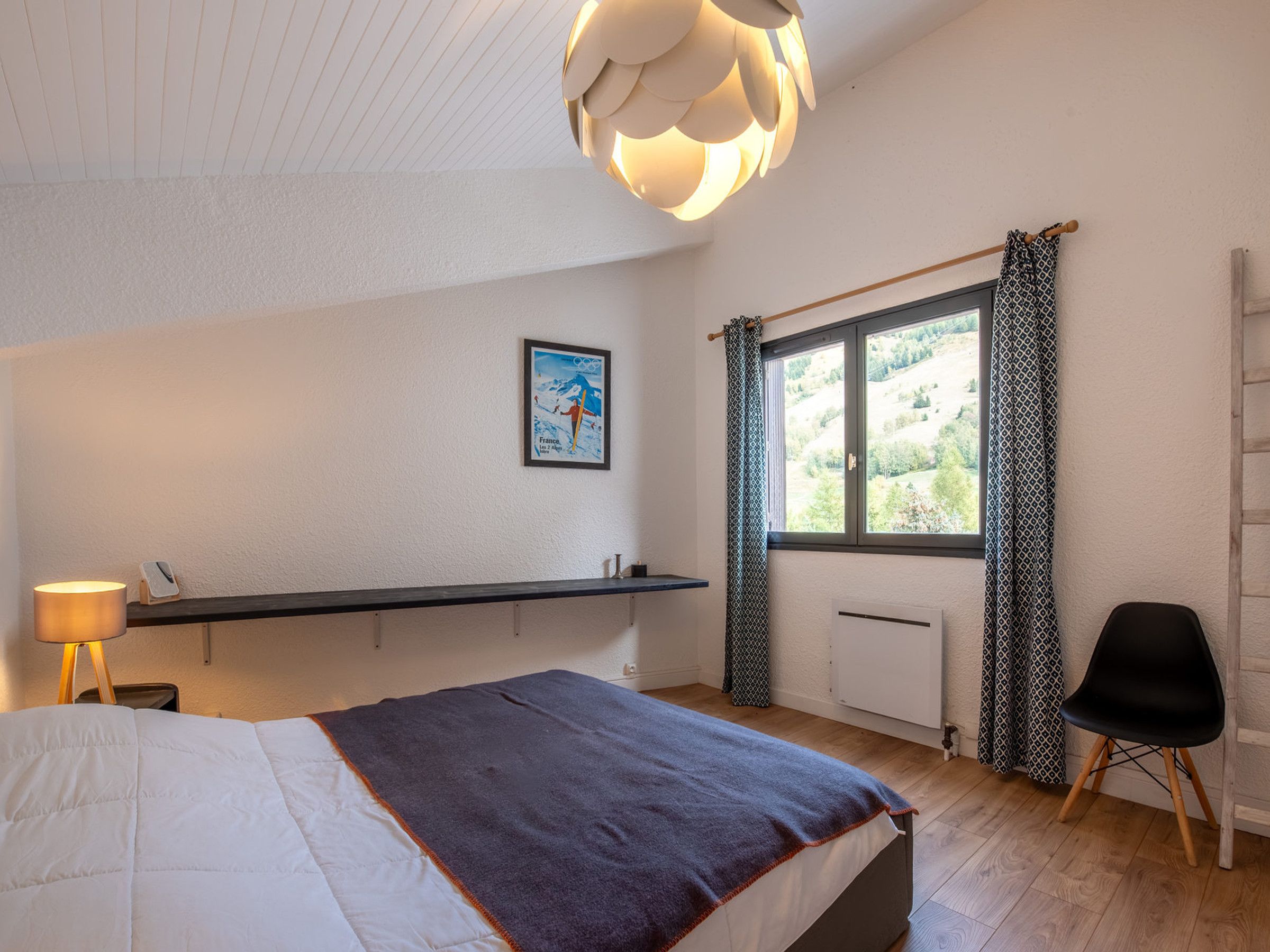 A bedroom with a bed, a bedside table, a lamp, a window with curtains, a radiator, and a chair.