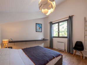 A bedroom with a bed, a bedside table, a lamp, a window with curtains, a radiator, and a chair.