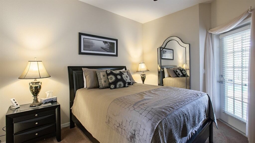 Une chambre à coucher avec un lit, deux tables de chevet avec des lampes, un tableau encadré au mur et une fenêtre avec des stores.