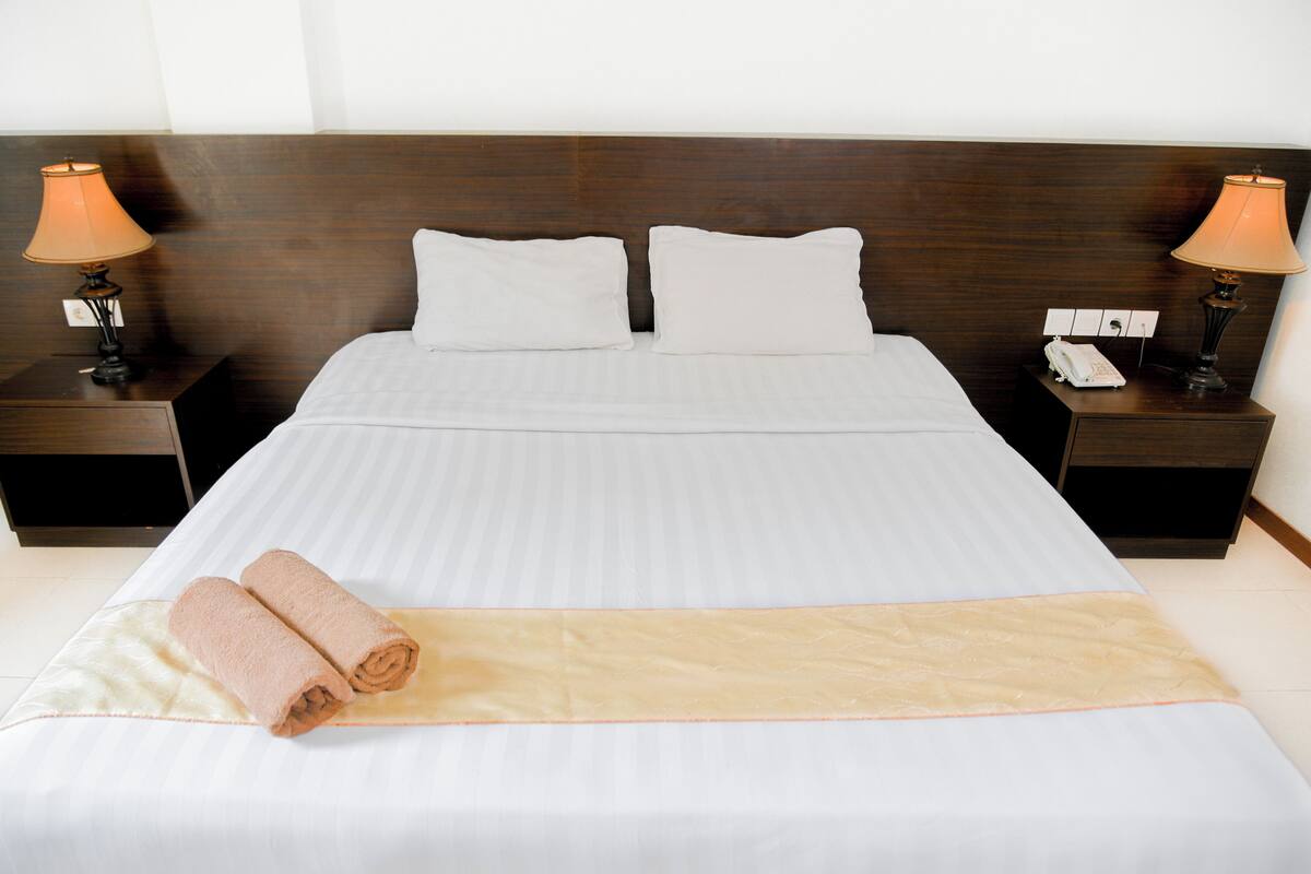 A neatly made bed with white linens, two pillows, and a beige bed runner. There are two bedside tables with lamps and a phone on one of them.