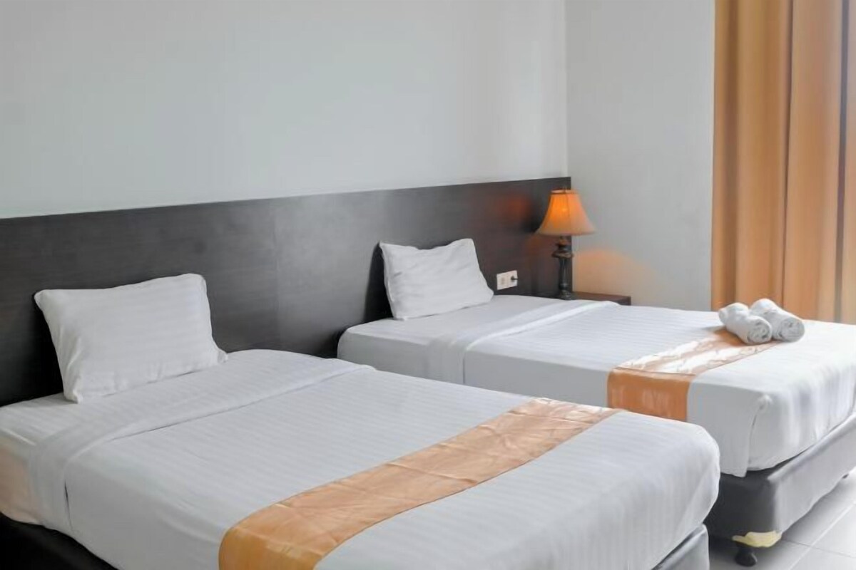 Habitación de hotel con dos camas individuales, una mesita de noche con lámpara y ventana con cortinas.