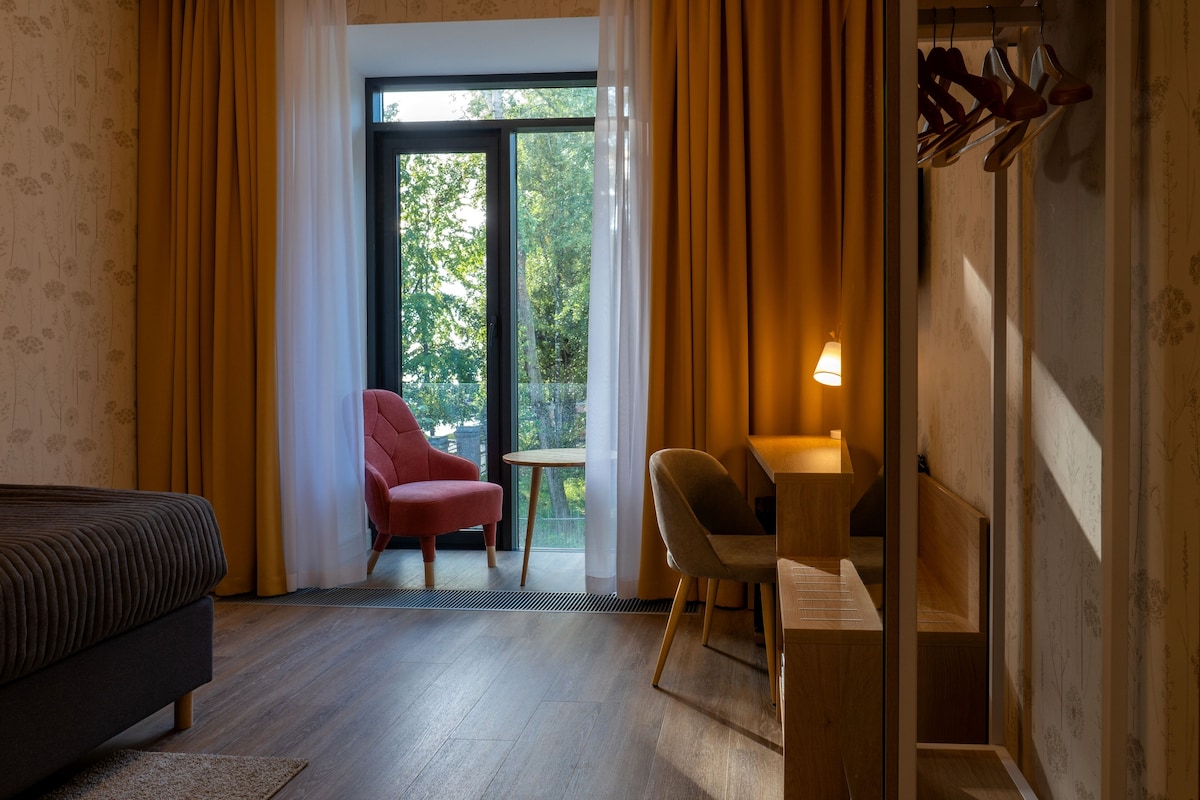 A hotel room with a bed, a red chair, a wooden desk, and a view through a large window.