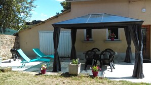 Ein überdachter Außenbereich mit Tisch, Stühlen, Blumentöpfen und einem Grill.