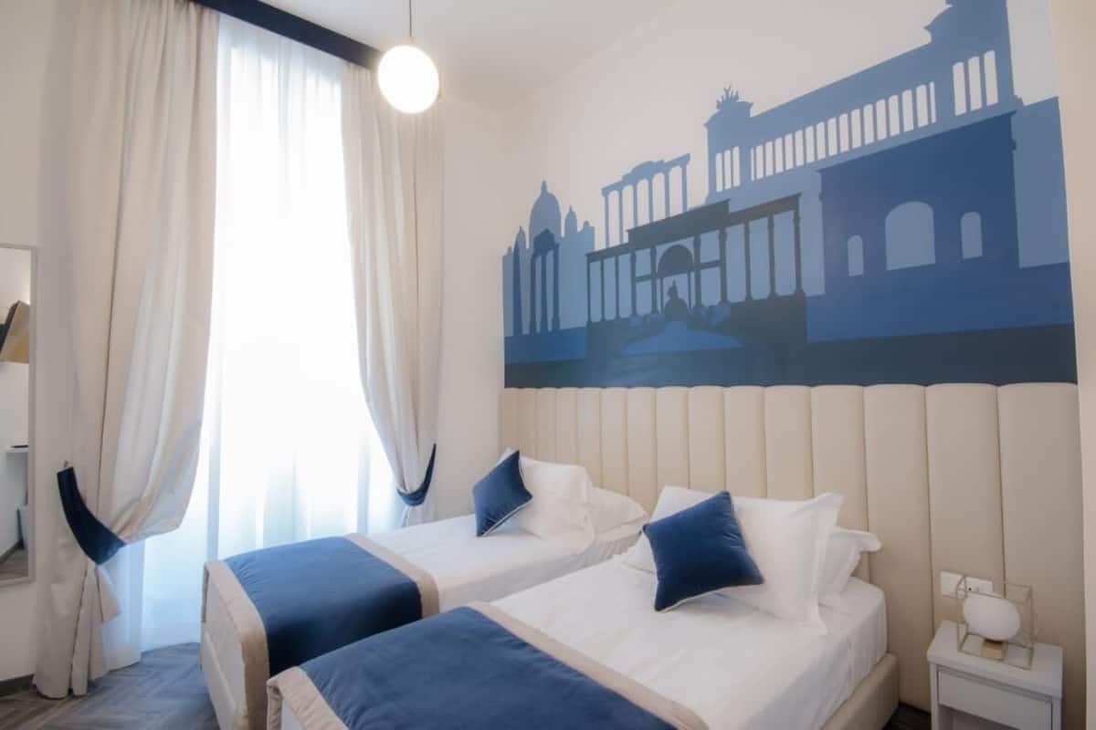 Habitación de hotel con dos camas, un ventanal con cortinas, un mural en la pared y una mesita de noche con lámpara.