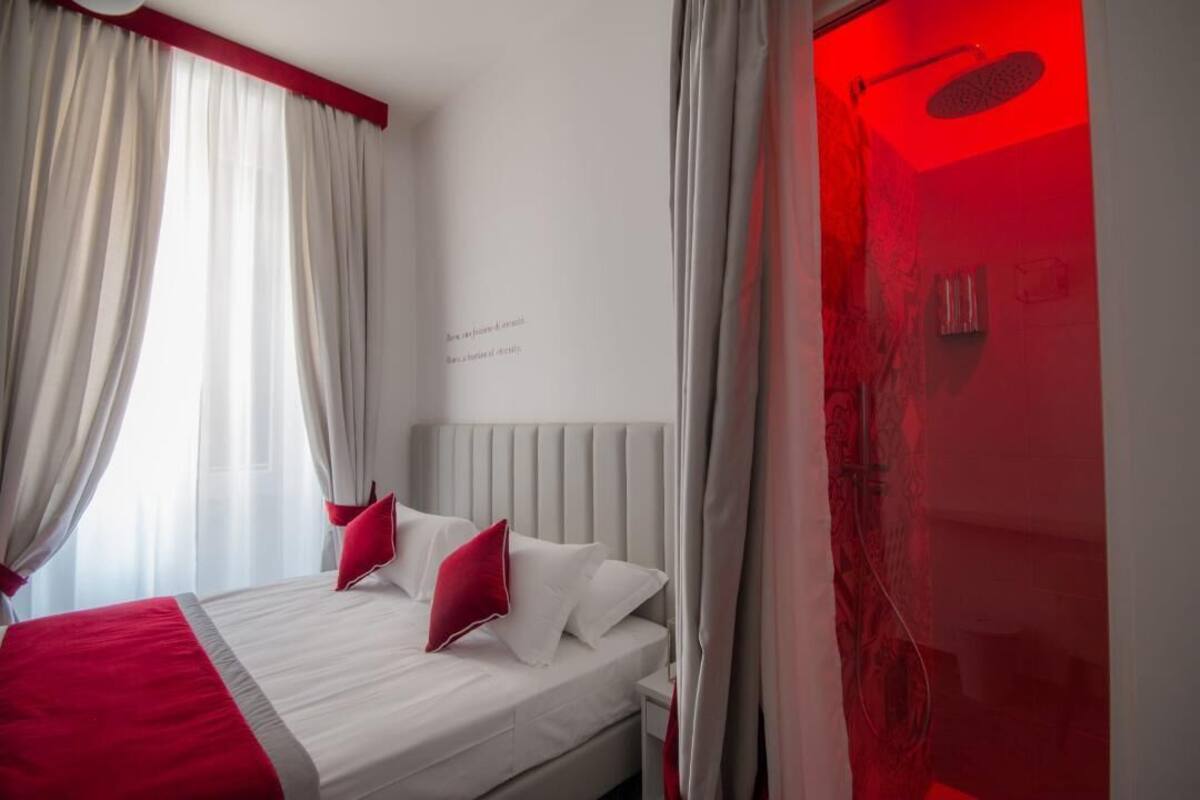 Habitación de hotel con cama, cortinas blancas, pared de acento roja y zona de ducha iluminada en rojo.