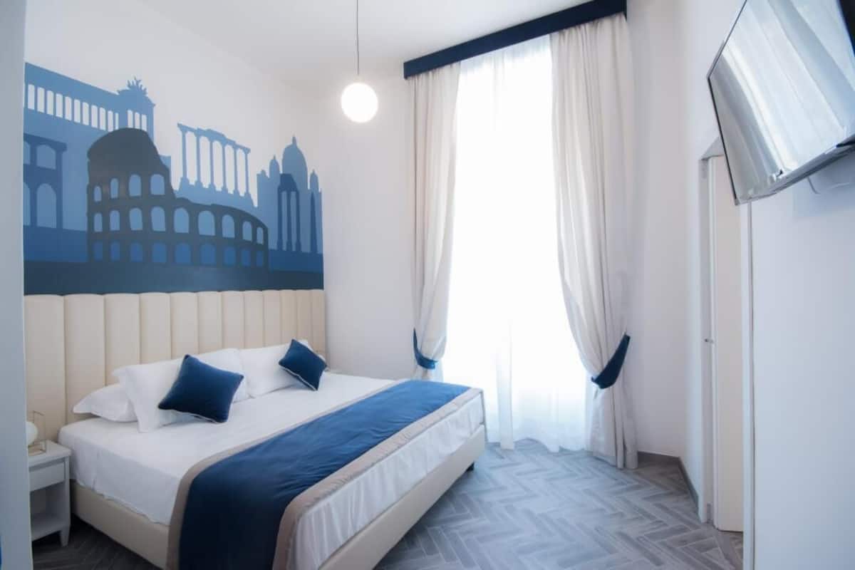 Una habitación de hotel con cama, un mural en la pared, televisor y ventana con cortinas.