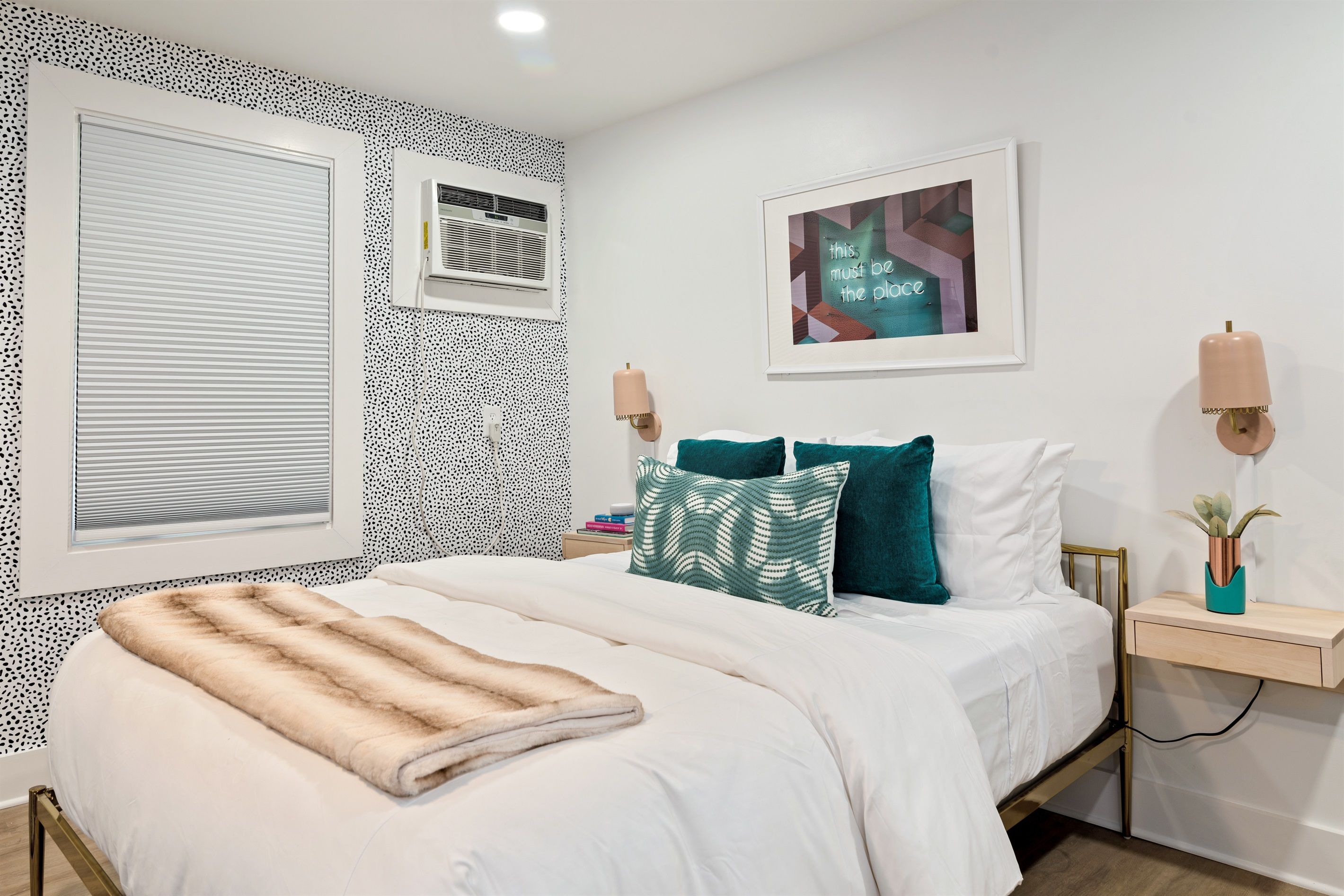 A bedroom with a bed, a window with blinds, a wall-mounted air conditioner, a framed artwork, two bedside lamps, and a bedside table with a plant.