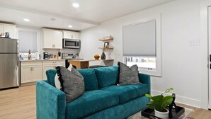 Ein modernes Wohnzimmer mit einem türkisfarbenen Sofa, einem Couchtisch mit Pflanze und einer Küche im Hintergrund.