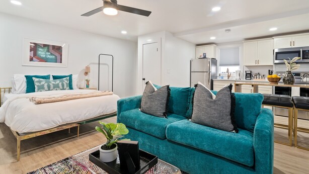 Ein modernes Wohnzimmer mit einem blaugrünen Sofa, einem Bett mit weißer Bettwäsche und einer Küche im Hintergrund.