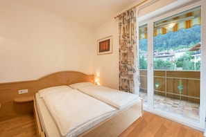 A bedroom with a bed, a nightstand, a wall-mounted light, a window with curtains, and a balcony with a view of a forested area. - Holiday Apartment "Residence Kronstein - 4" with Balcony, Shared Garden and Mountain View (Tisens Italien)