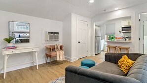 Una sala de estar moderna con un sofá gris, una mesita y un escritorio. Hay una puerta que da a una zona de cocina con un refrigerador y una ventana.