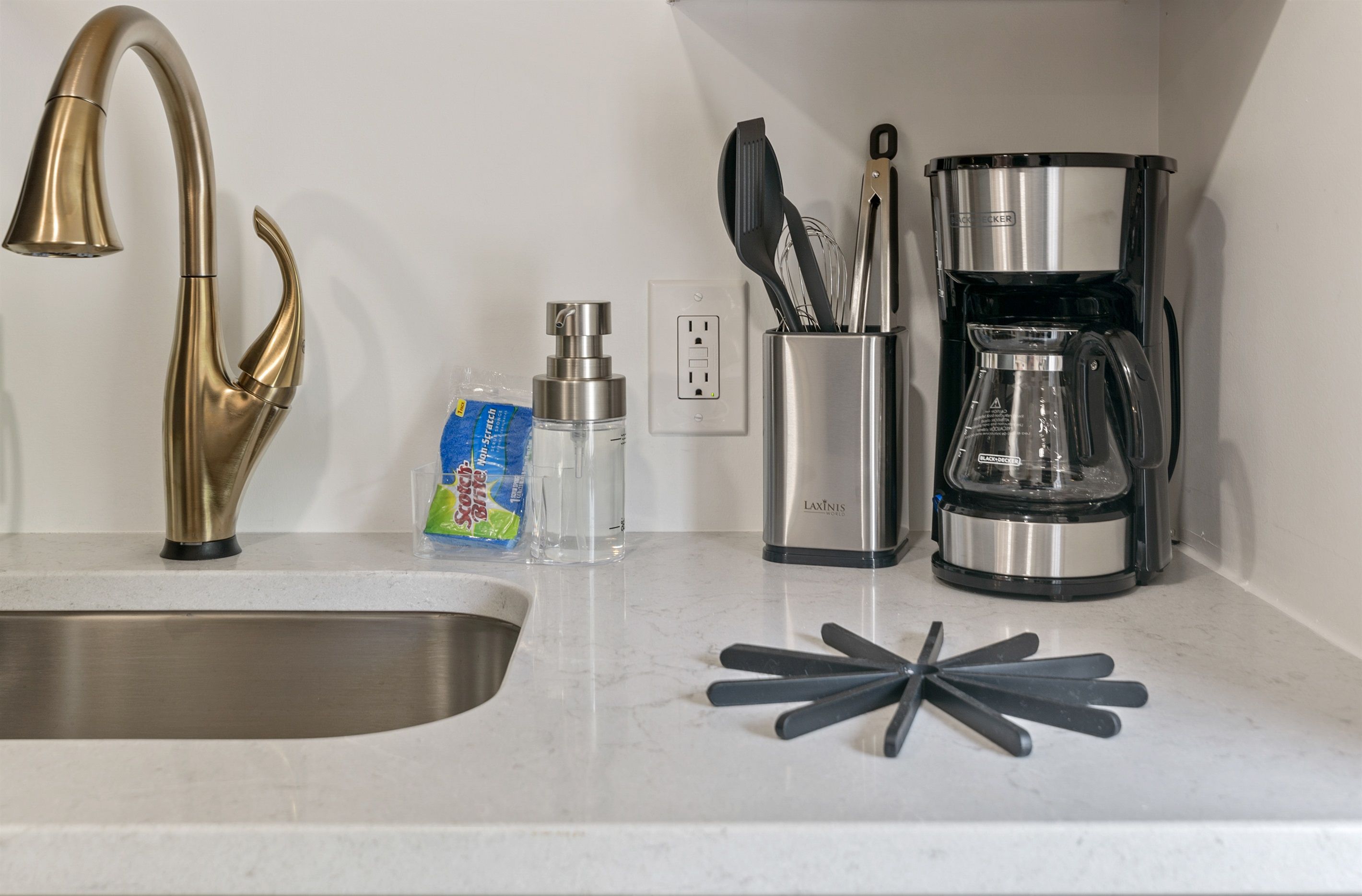 Una encimera de cocina con un lavamanos de acero inoxidable, un grifo dorado, una cafetera y utensilios de cocina.