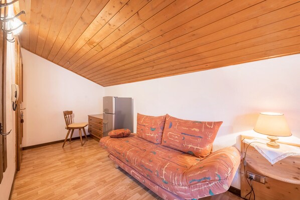 A room with a wooden ceiling, a sofa with patterned cushions, a chair, a small table with a lamp, and a refrigerator. - Apartment "Residenz Kronstein - 7" with Balcony, Shared Garden and Mountain View (Tisens Italien)
