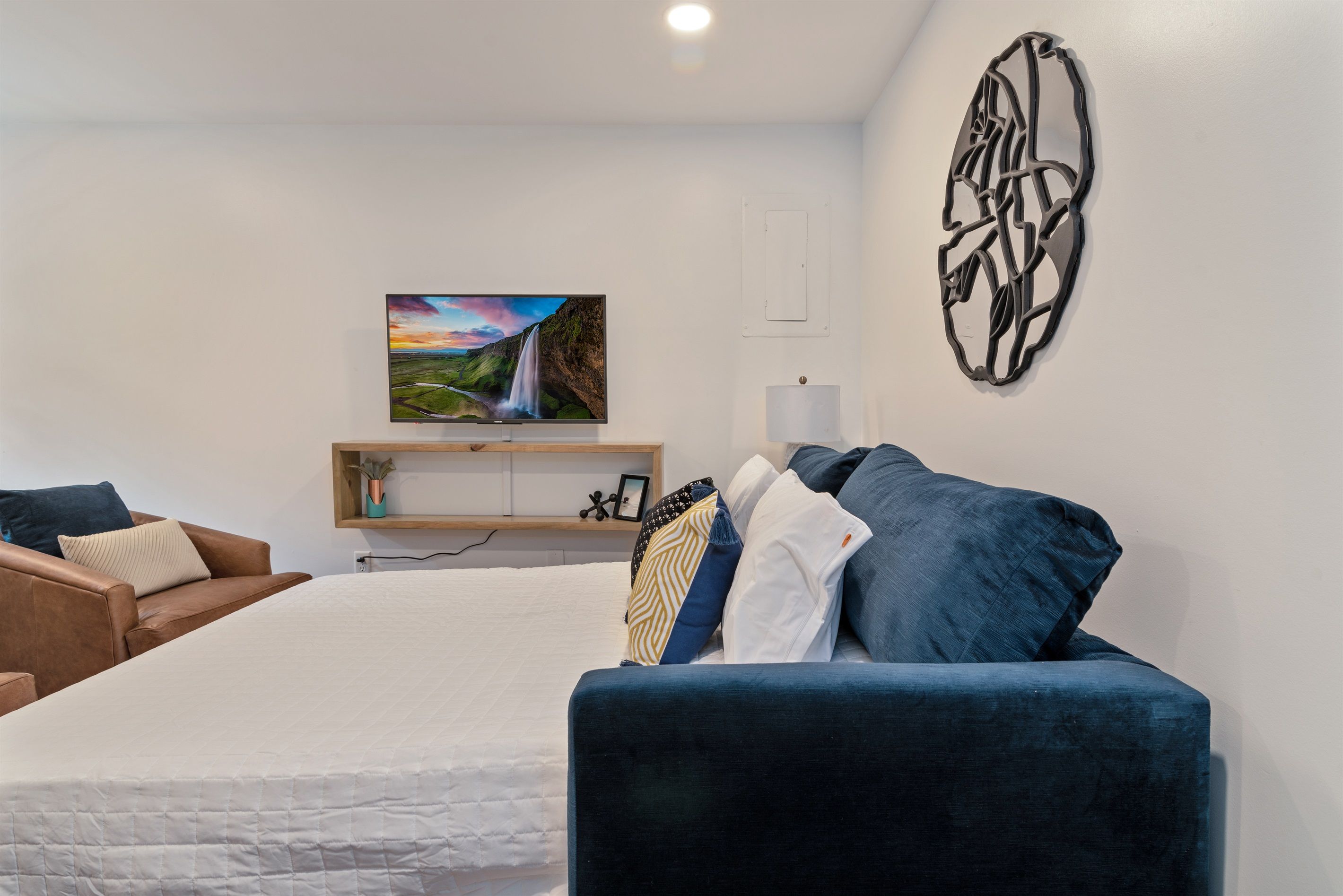A bedroom with a bed, a blue sofa, a TV on a shelf, and a wall art piece.