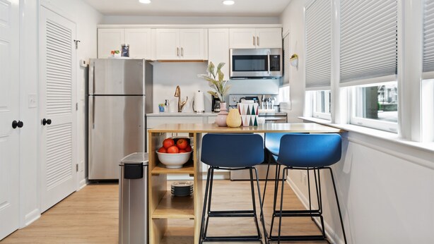 A modern kitchen with white cabinets, a stainless steel refrigerator, a microwave, and a sink. There are two blue bar stools and a wooden shelf with fruits.