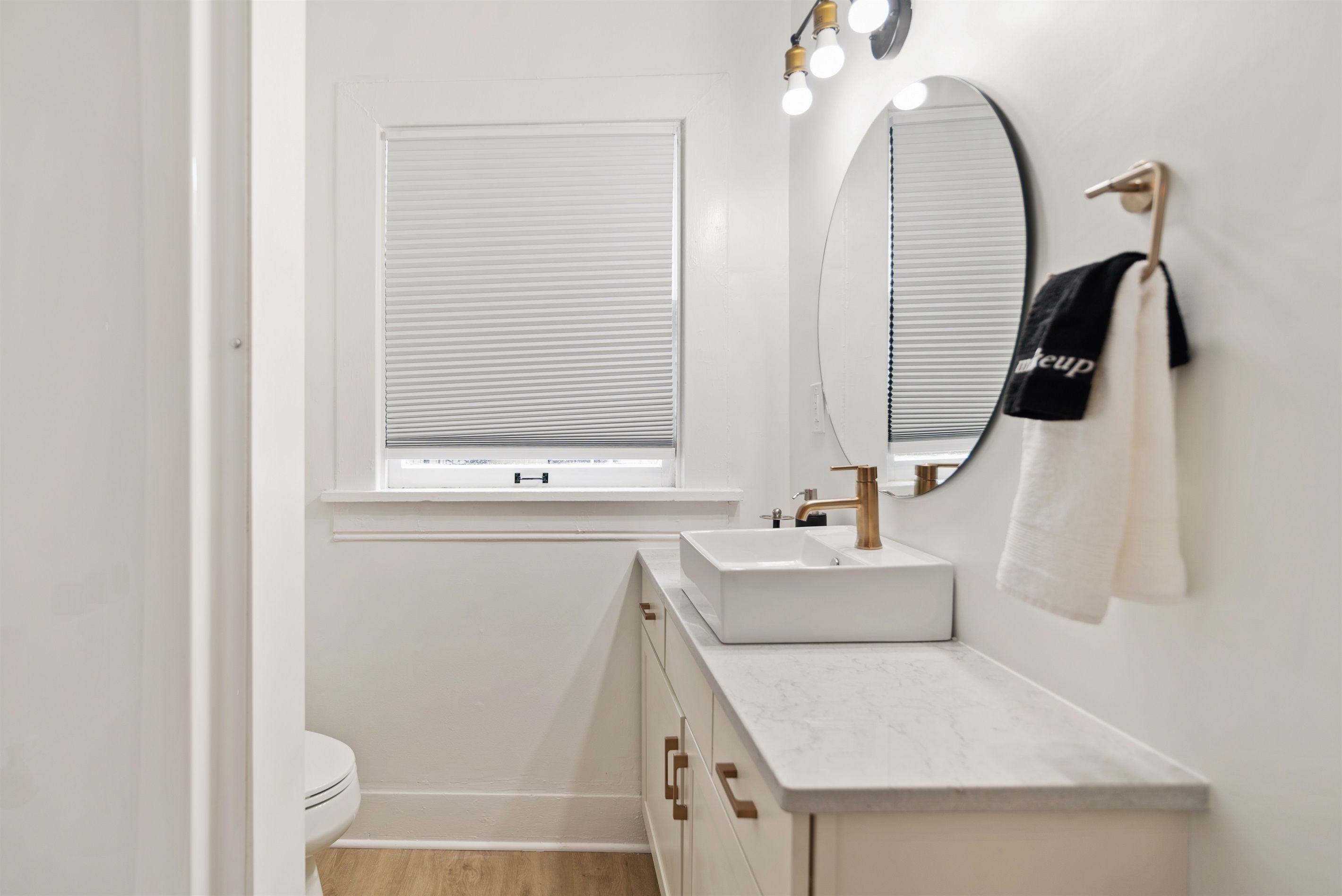 A modern bathroom with a white sink, a mirror, and a window with blinds.