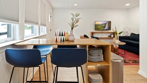 A modern living room with a dining area, featuring a wooden table, blue chairs, a vase with plants, and a painting on the wall.