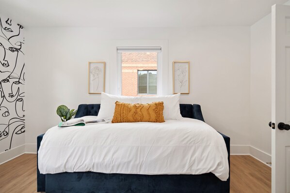 A bedroom with a dark blue bed, white bedding, a window with a view of a brick building, and two framed artworks on the wall.