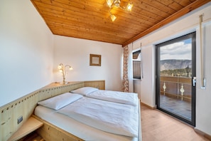 Une chambre avec un plafond en bois, un lit avec une literie blanche, une table de chevet avec une lampe, et une vue à travers une porte vitrée.