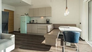 Una cocina moderna con horno empotrado, un comedor con mesa blanca y sillas azules, y una zona de estar con sofá y banco.