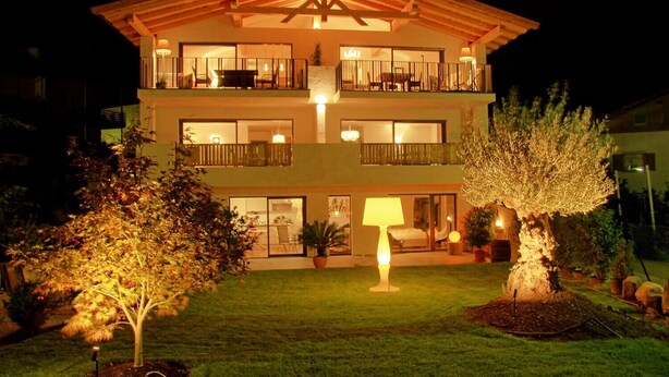 A multi-story building with balconies, a well-lit entrance, and a landscaped garden with trees.