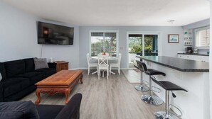 A modern living room with a black sofa, a wooden coffee table, and a TV mounted on the wall. There is a dining area with a white table and chairs, and a kitchen area with a bar counter and stools. - #155 Nestucca Sunrise · Riverfront, Close to Ocean Beaches, Large Yard (Pacific CIty)